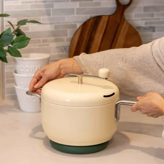 Person holding a cream-colored pot with a lid on a kitchen counter.