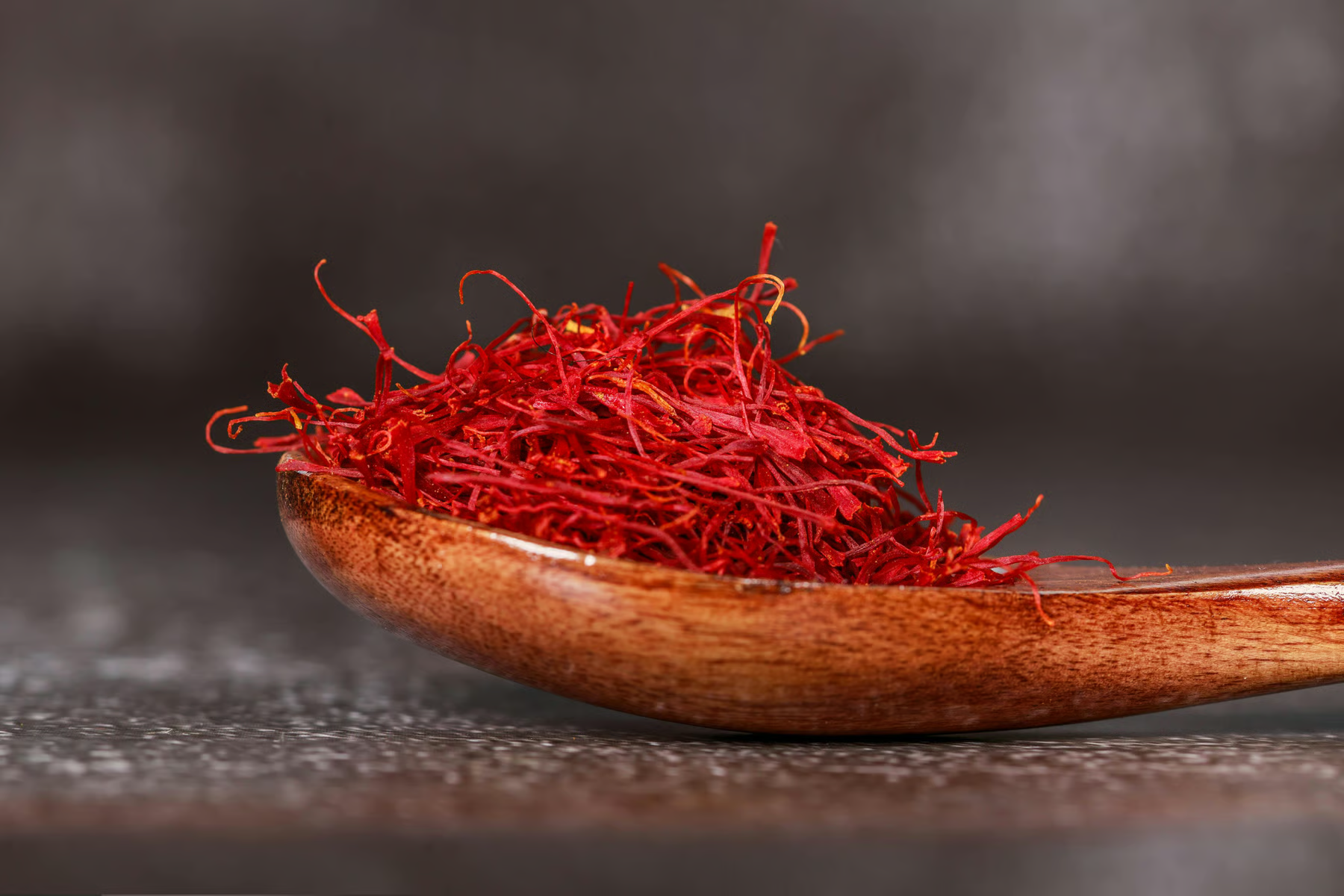 A close-up of a wooden spoon overflowing with bright red saffron threads against a dark background.