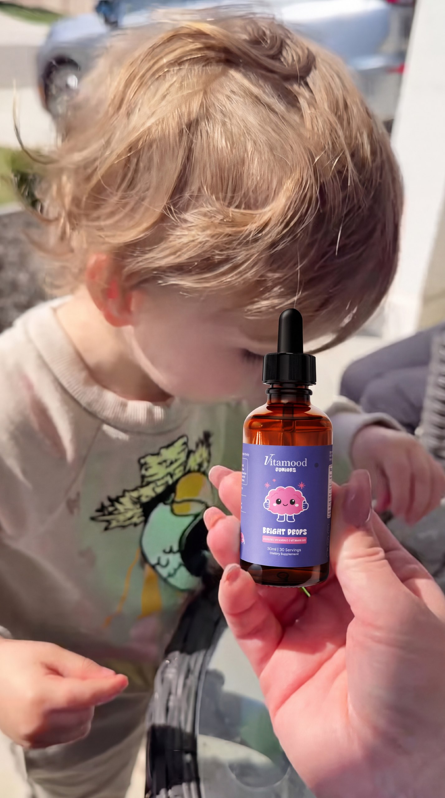 A hand holding a dropper bottle of Vitamood Juniors Bright Drops with a toddler in the background.