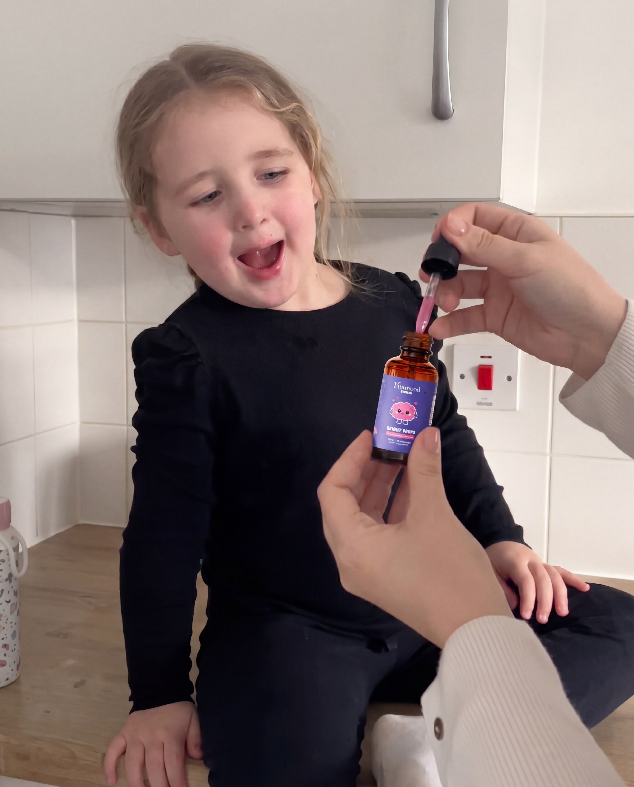 A young girl sits on a counter, looking excitedly at an adult holding a bottle and a dropper.