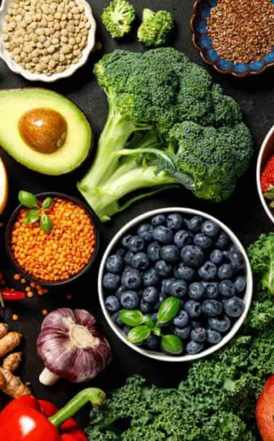 An overhead shot of fresh vegetables, fruits, legumes, and seeds arranged on a dark surface.