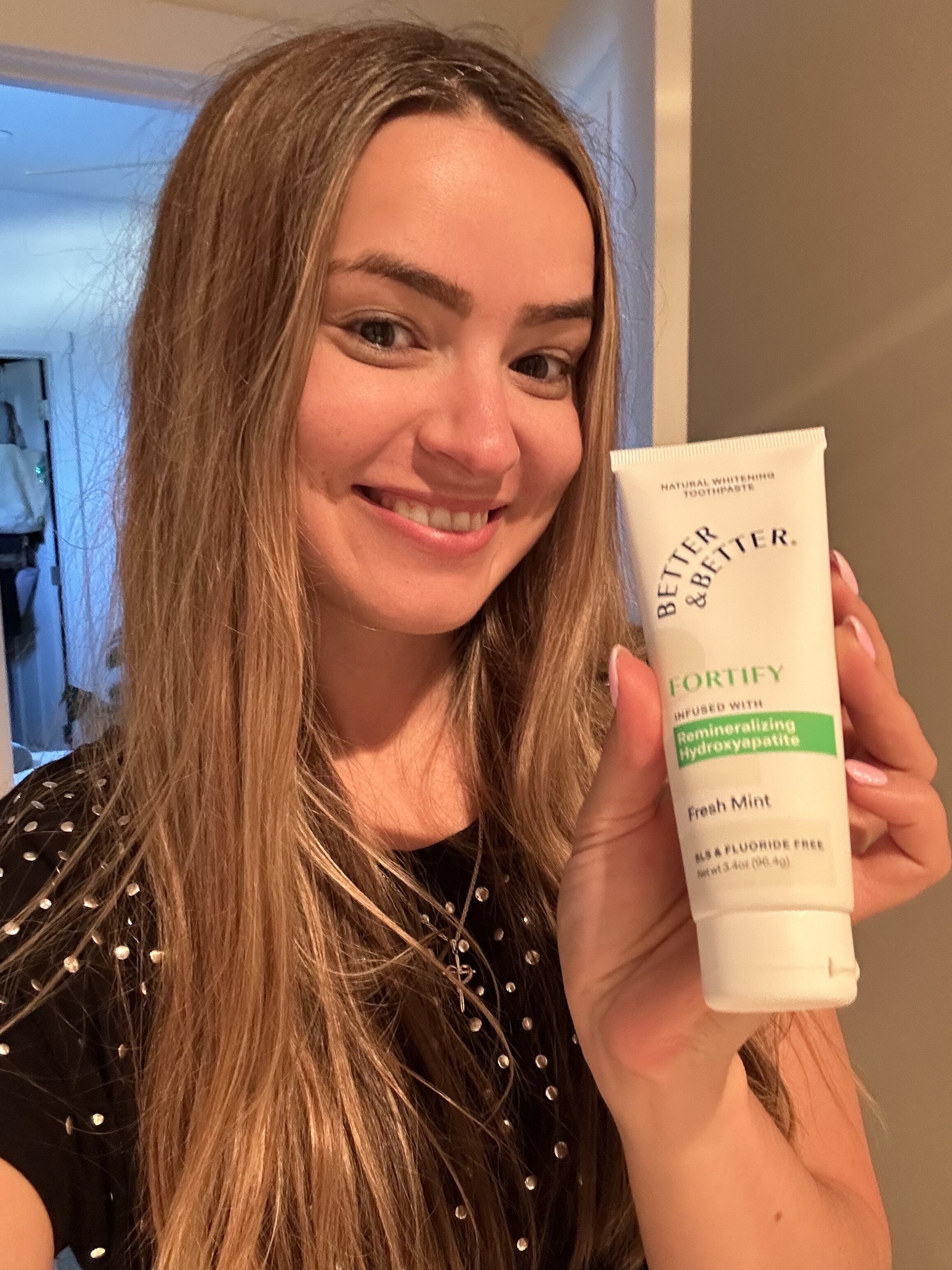Person smiling and holding a tube of toothpaste labeled 'Better & Better Fortify Fresh Mint.'