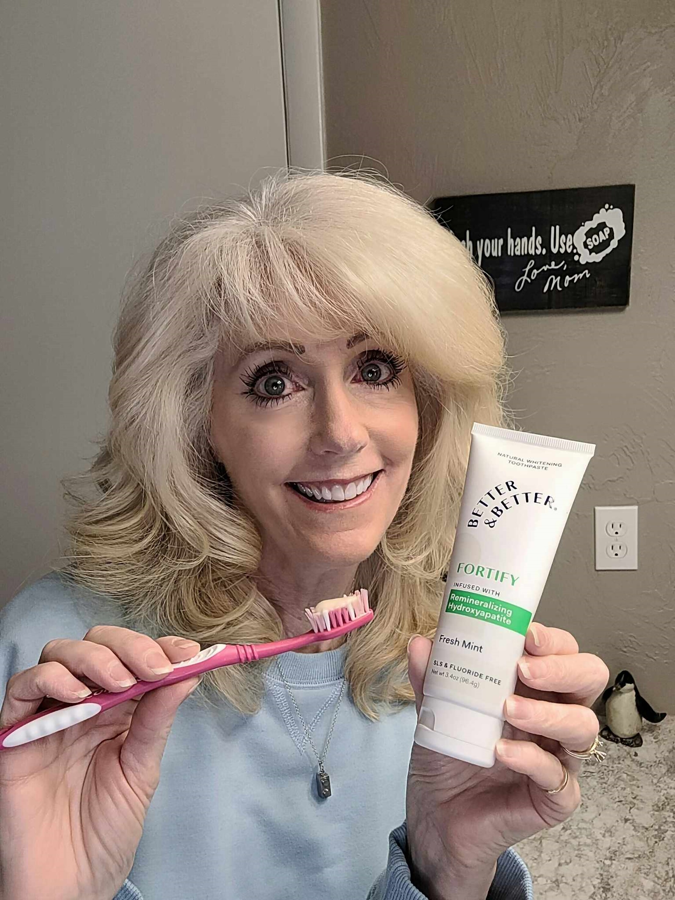 Person holding a toothbrush and toothpaste, smiling in a bathroom.