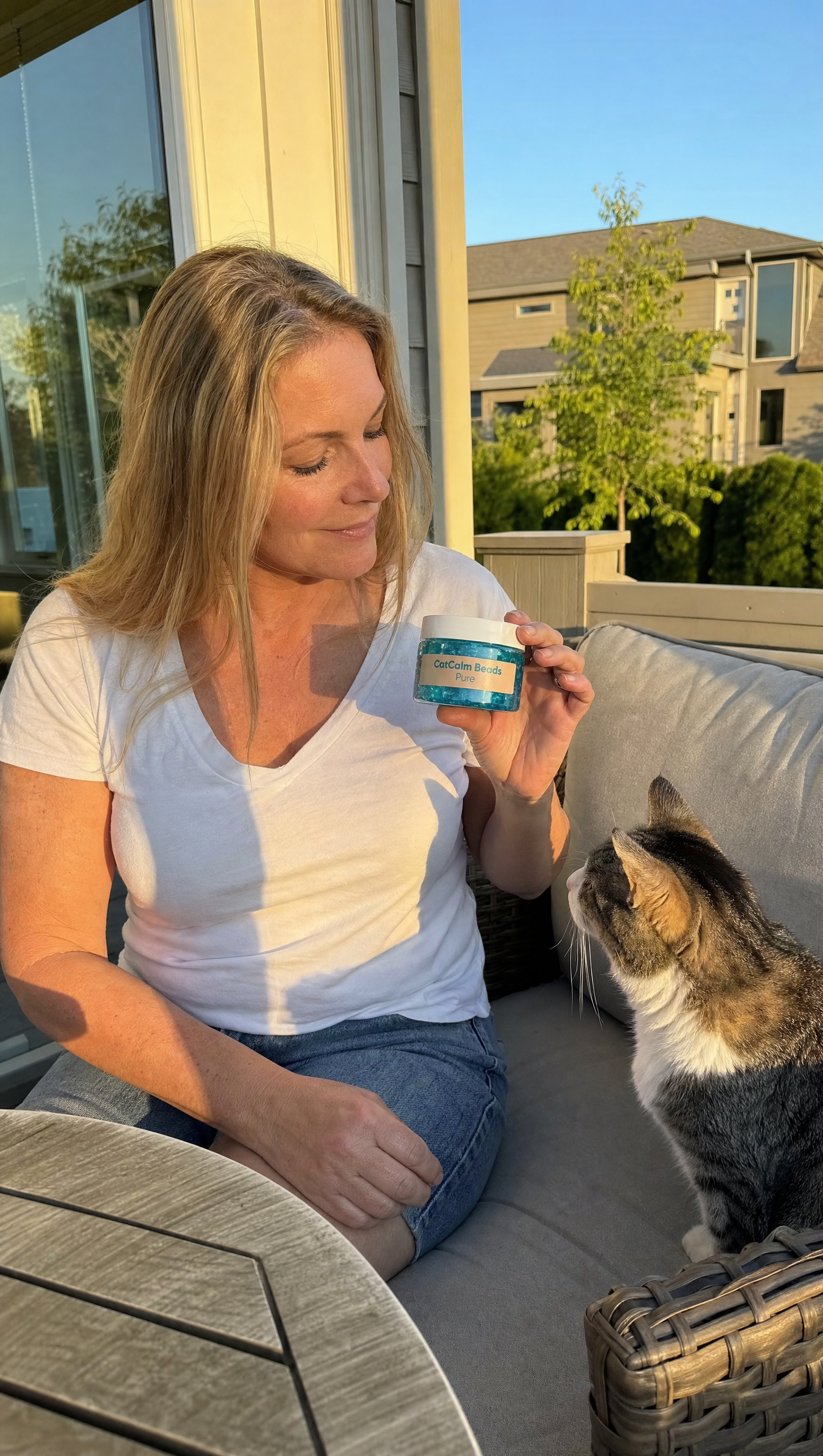 A woman on a patio holds a small jar of 'CatCalm Beads' as a cat looks up at it.