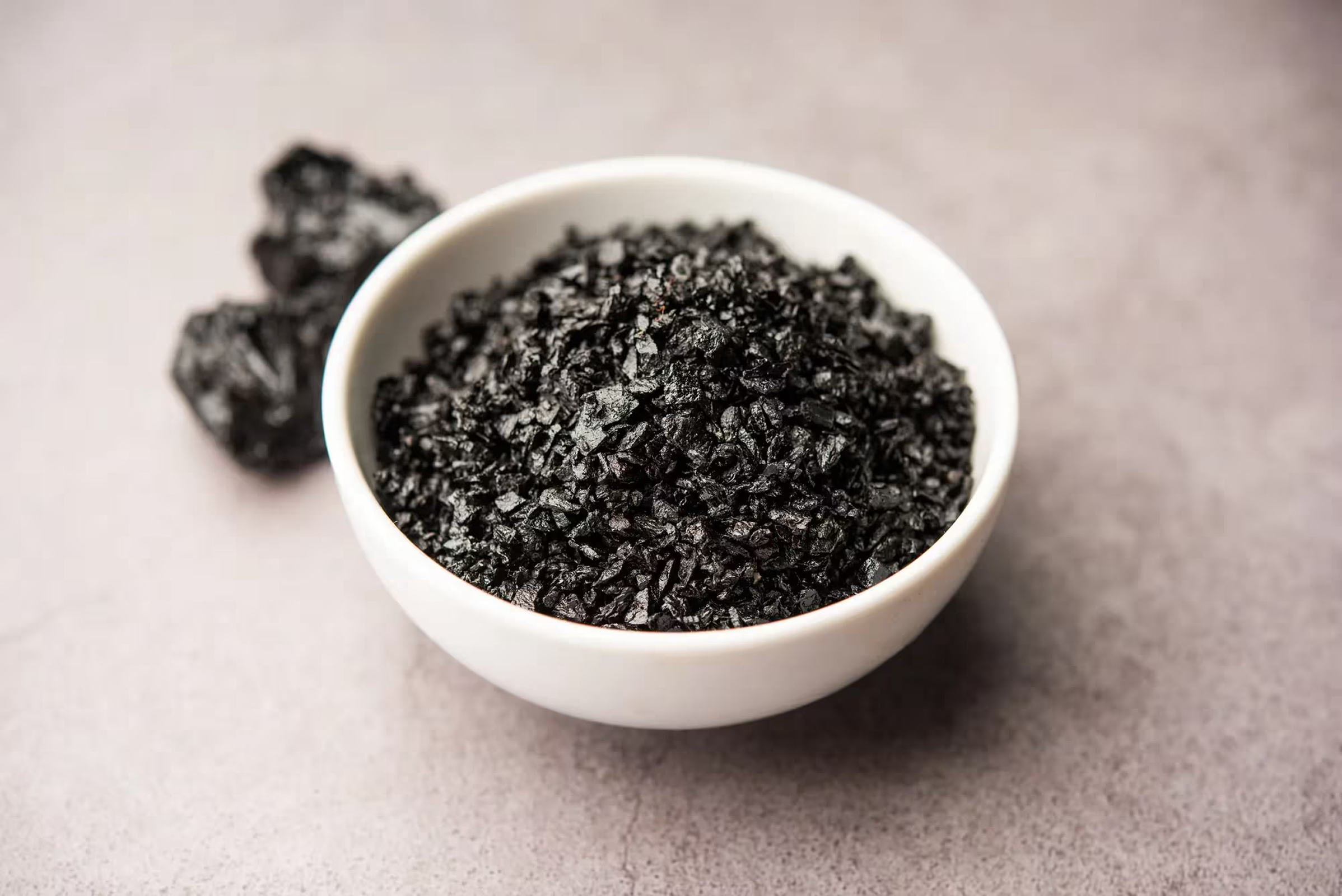 A small white bowl filled with coarse black salt flakes, with larger salt chunks in the background.