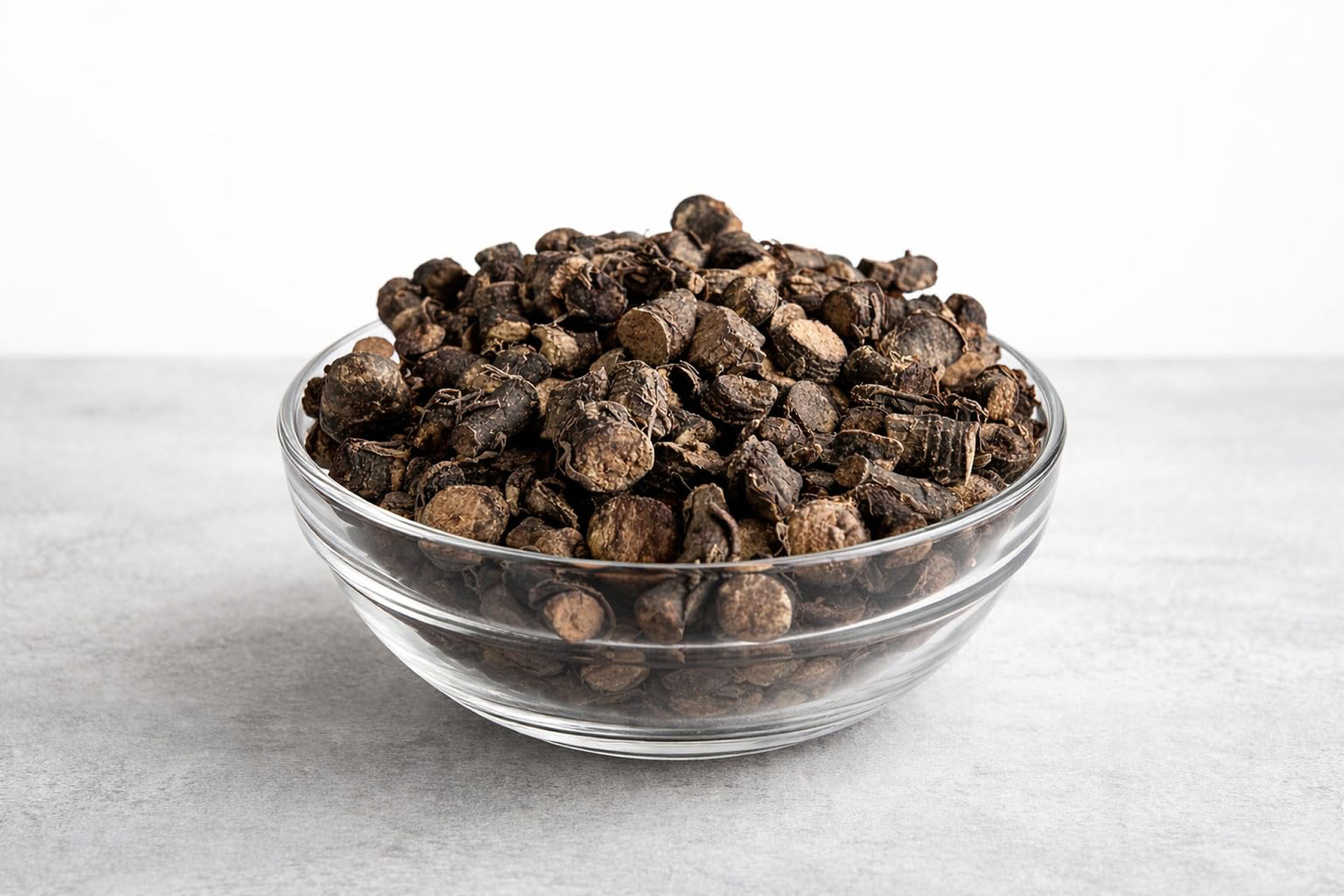 A clear glass bowl filled with pieces of dried, dark root on a grey surface against a white background.