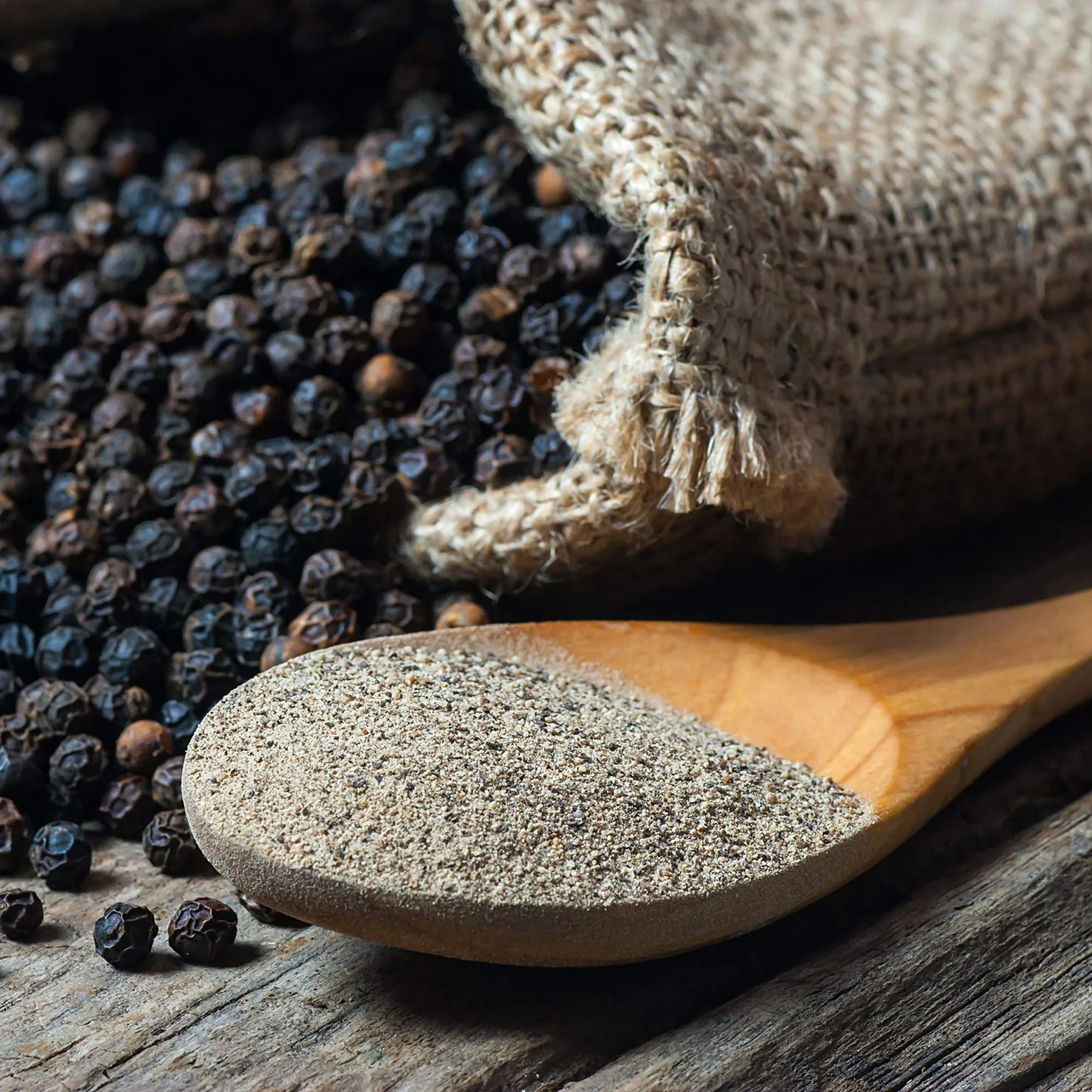 A wooden spoon of ground pepper sits before a burlap sack spilling whole black peppercorns onto wood.