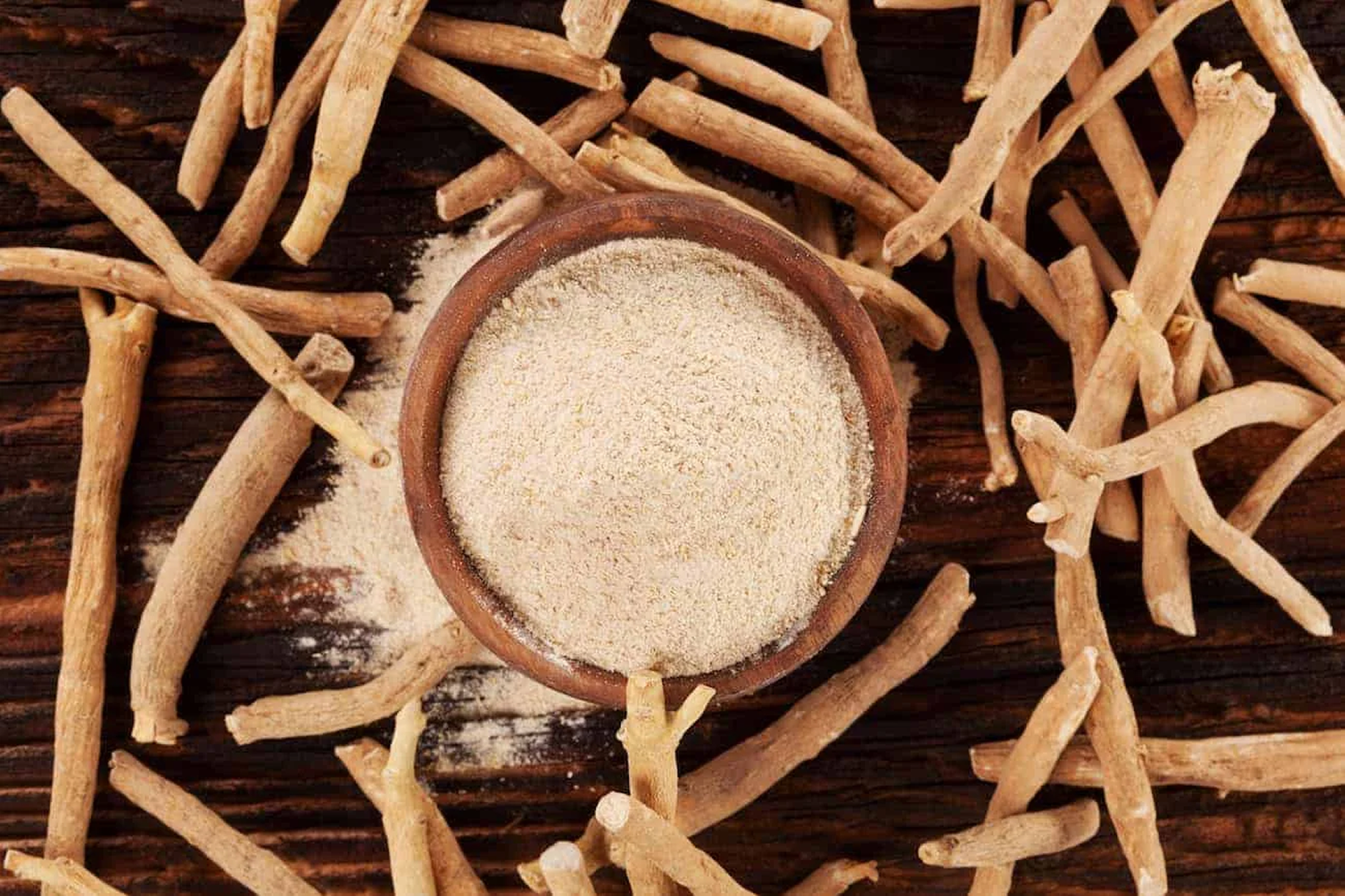 An overhead shot of a wooden bowl of powder surrounded by dried roots on a wooden table.