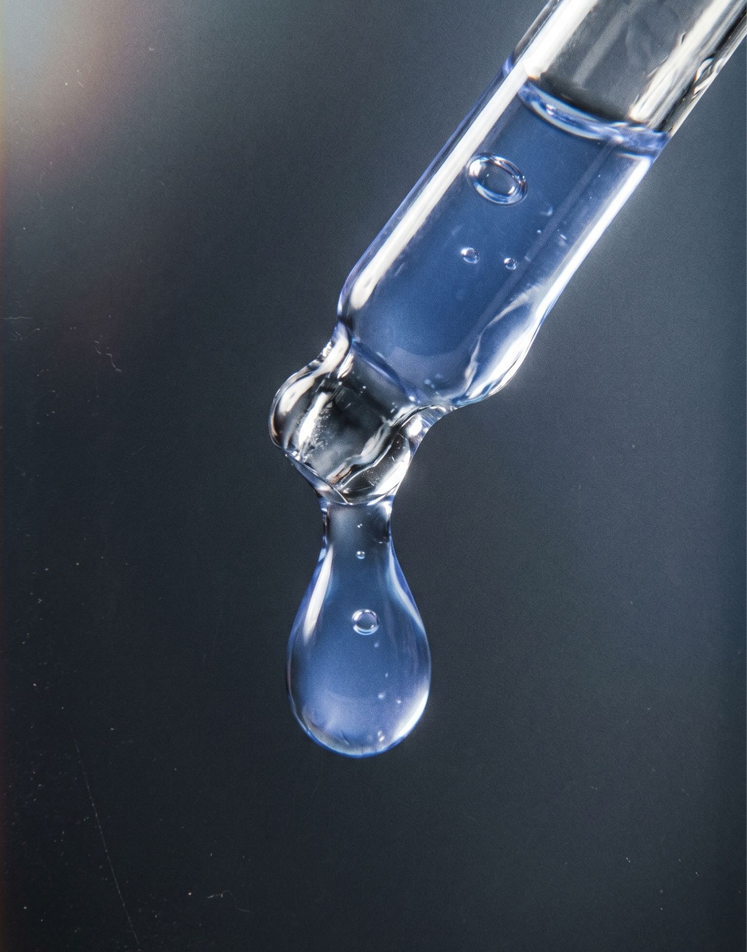 A close-up of a drop of blue liquid falling from a glass dropper against a dark background.