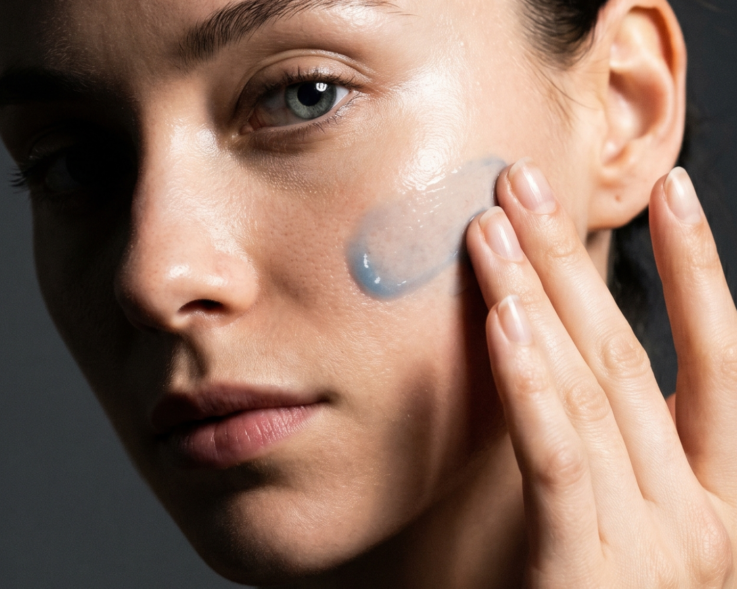 A close-up of a woman applying a translucent blue gel to her cheek with her fingertips.