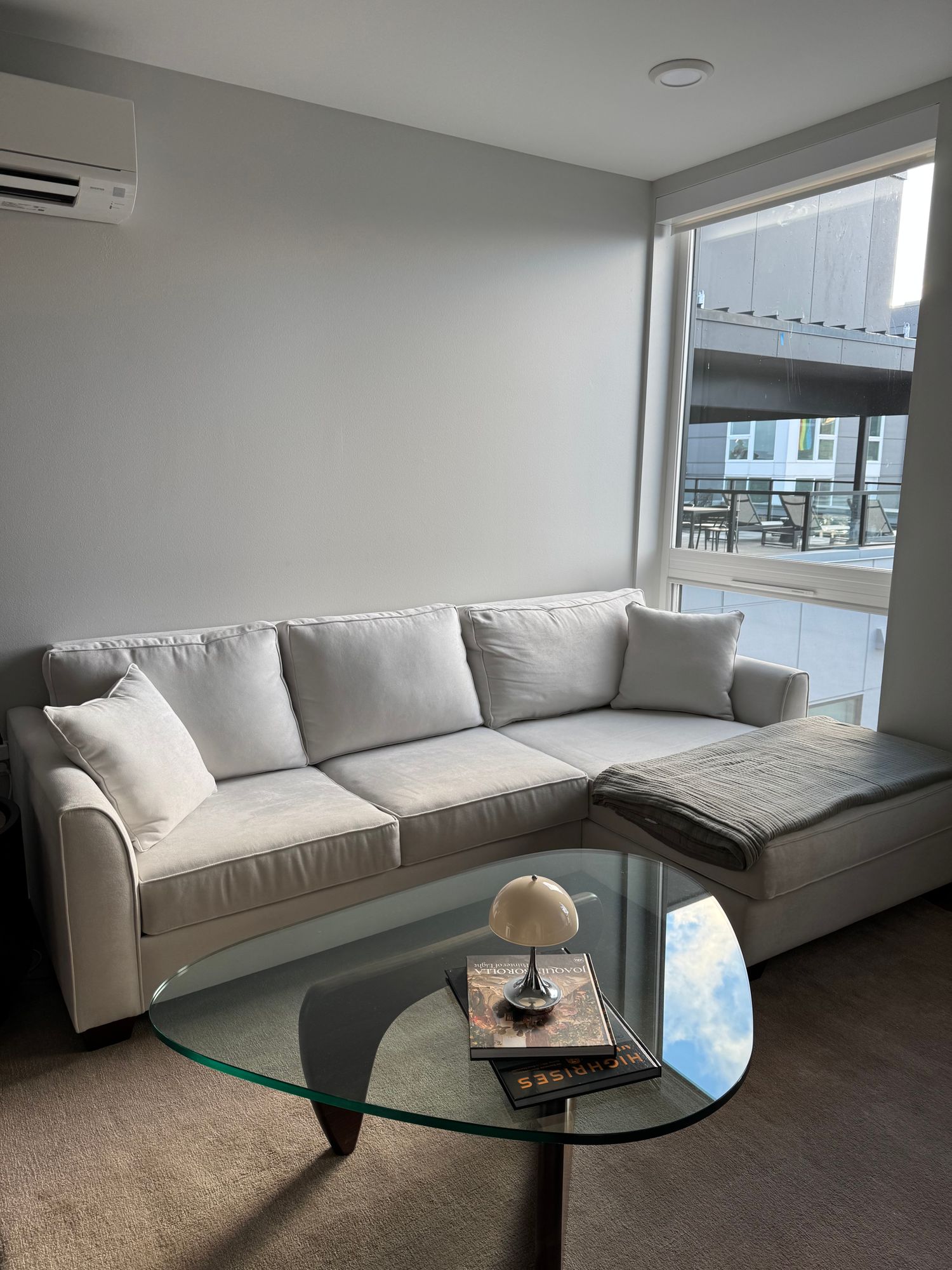 A modern living room with a light-colored sectional sofa, a glass coffee table, and a large window.