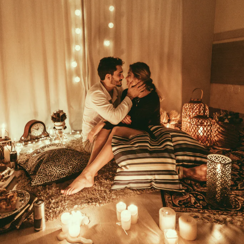 A couple embraces on the floor in a romantic room illuminated by candles and string lights.