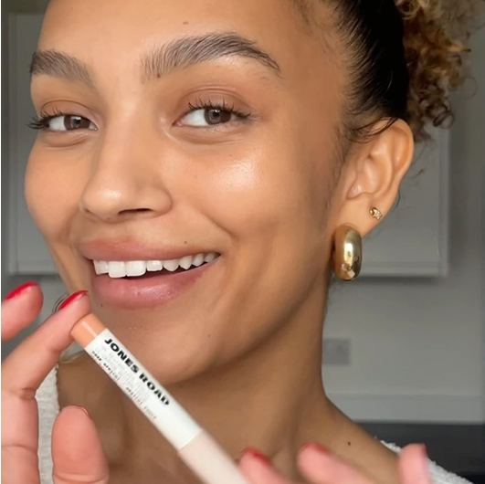 Person smiling, holding a Jones Road makeup product, with a natural look.