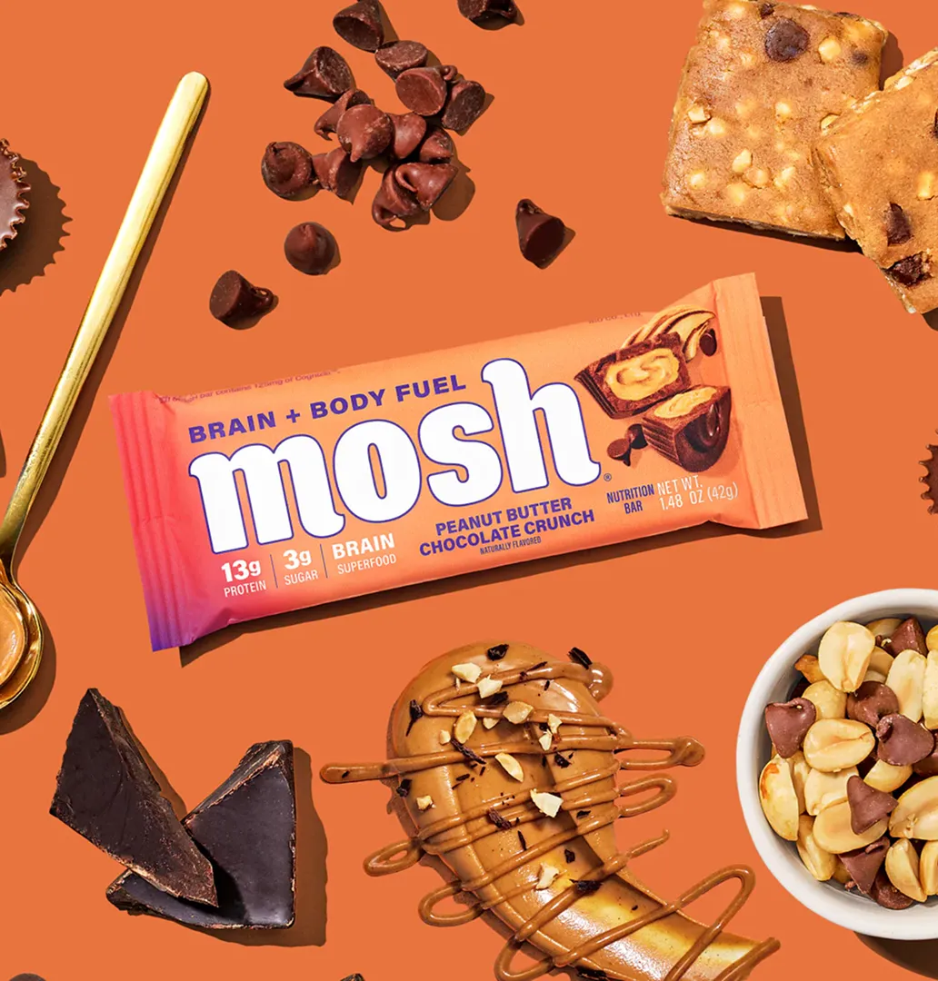 A Mosh protein bar on an orange surface, surrounded by peanuts, chocolate chips, and peanut butter.