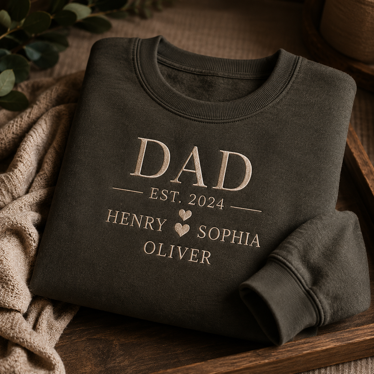 A dark gray sweatshirt embroidered with 'DAD EST. 2024' and three children's names folded in a wooden tray.