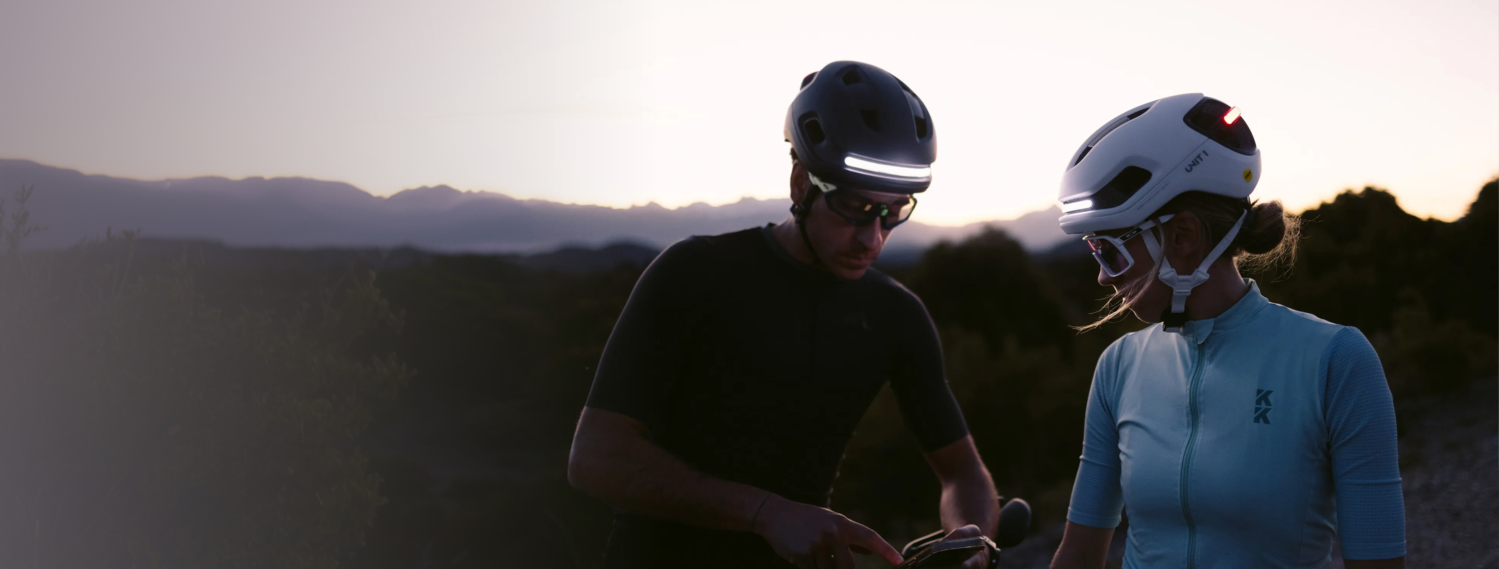 Two cyclists wearing helmets with built-in lights stand with their bikes at dusk.