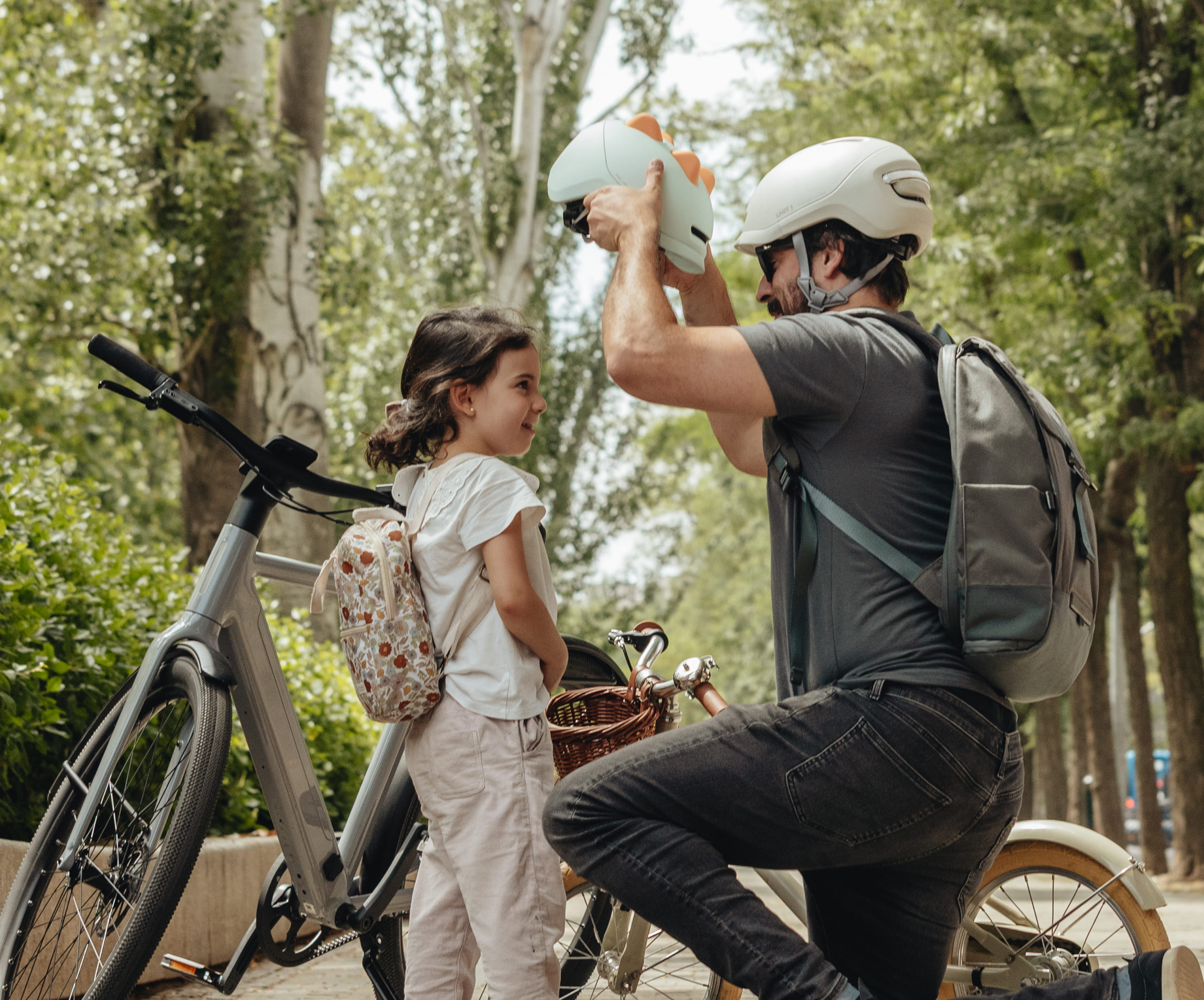 Adult helping child with a helmet near bicycles in a forested area.
