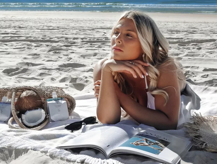 Woman relaxing on a beach with a magazine, coconut, and beach bag.