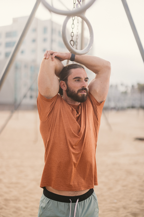 Man in orange shirt stretching under gymnastic rings on a sandy beach.