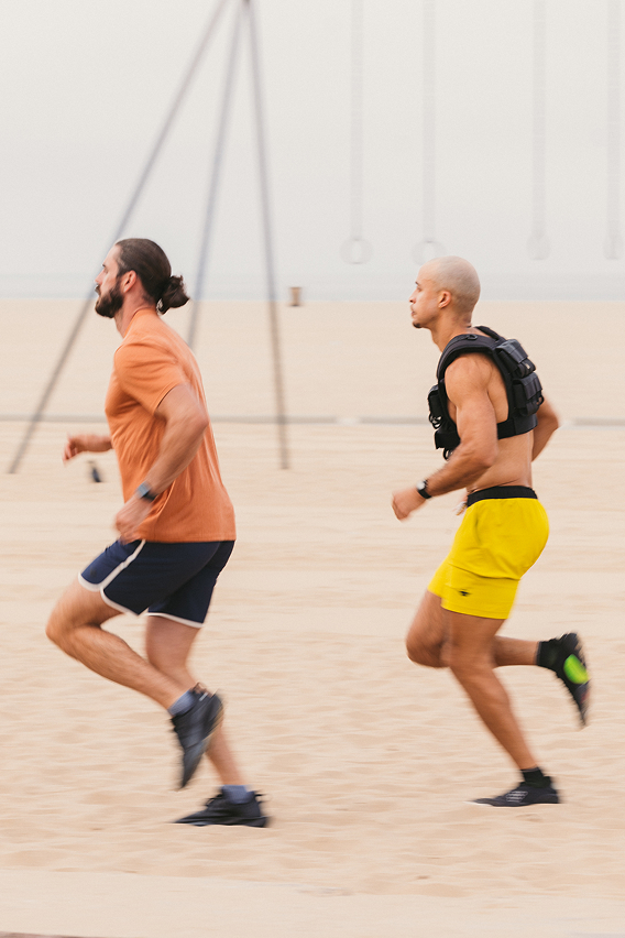Two men running on a sandy beach, one wearing a weighted vest.