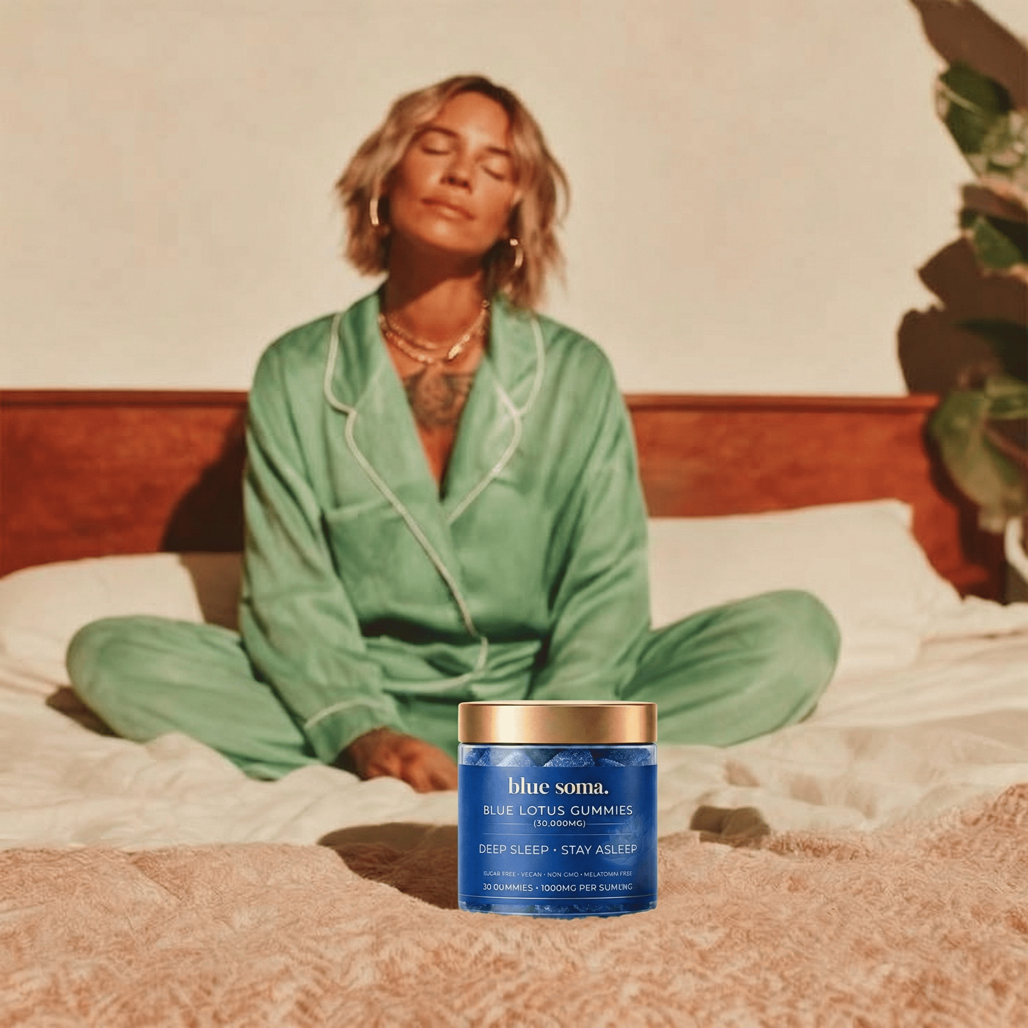 A woman in green pajamas sits relaxed on a bed with a jar of Blue Soma gummies.