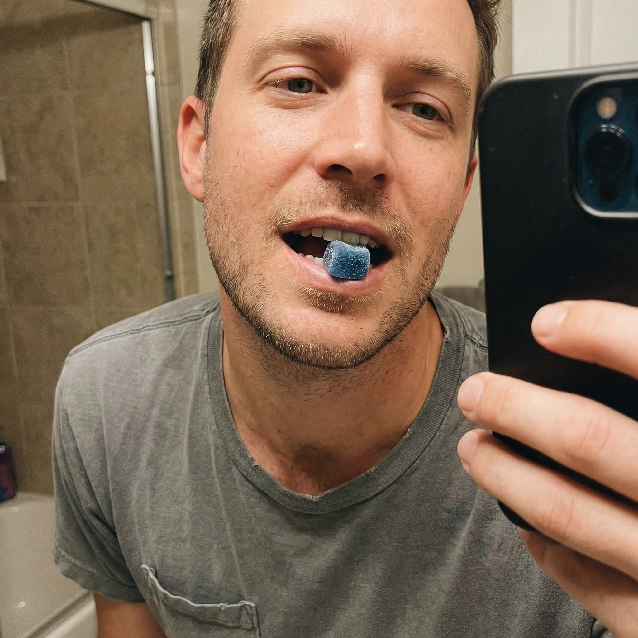 A man in a gray t-shirt takes a selfie with a blue, sugar-coated gummy in his mouth.