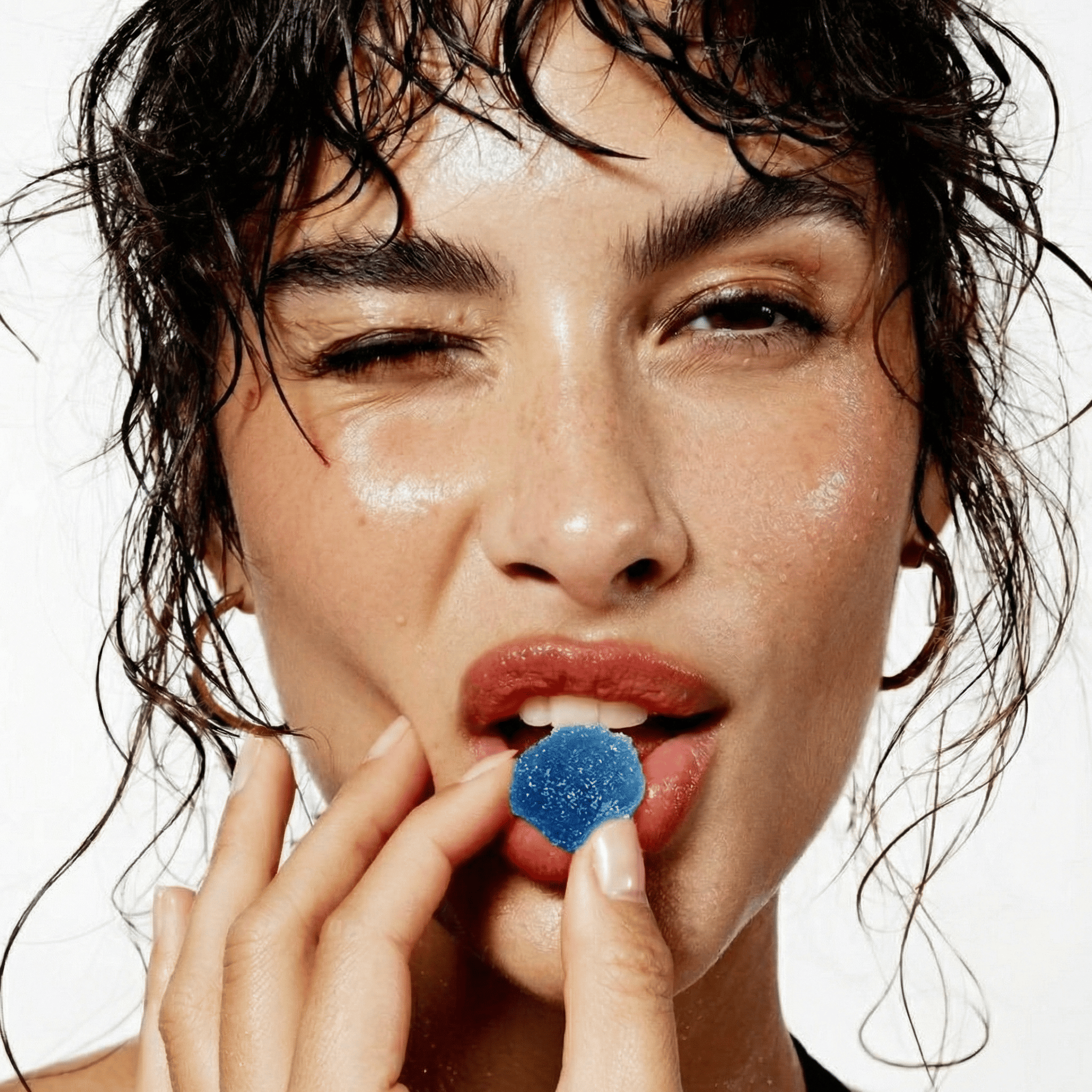 A woman with wet hair winks while holding a blue gummy to her lips against a white background.