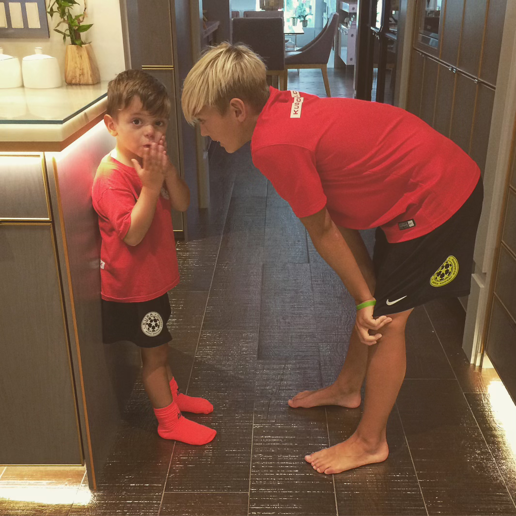 Two children in red shirts, one whispering to the other in a hallway.