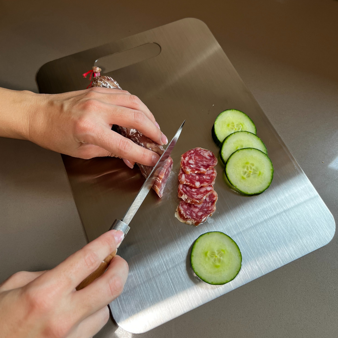 A person slices salami on a metal cutting board next to several slices of cucumber.