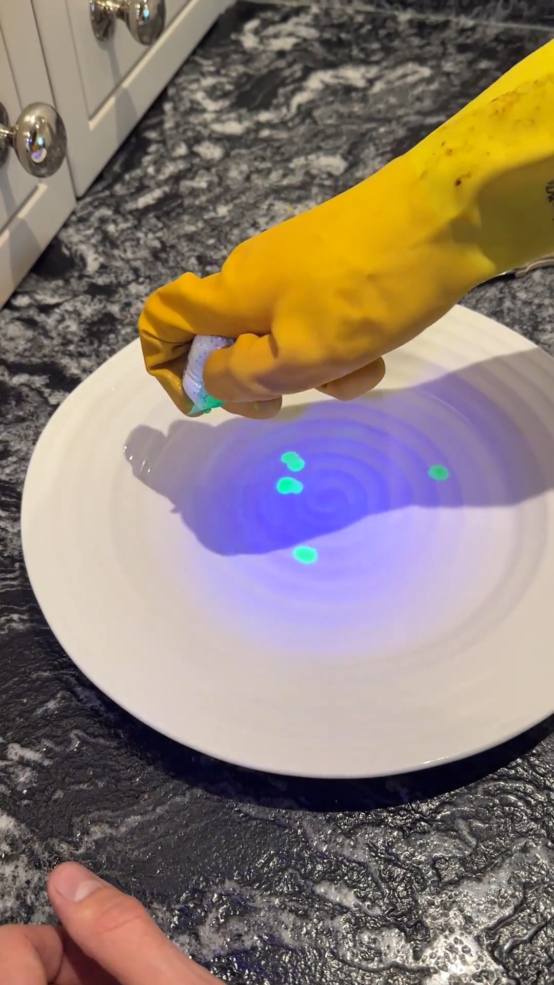 Yellow gloved hand applying green liquid to a white plate on a counter.