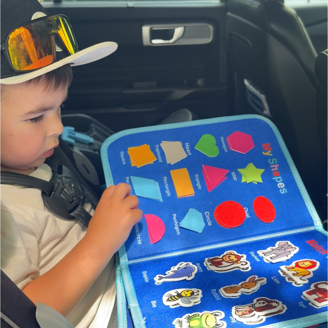A young child in a car seat plays with a blue felt activity book about shapes.