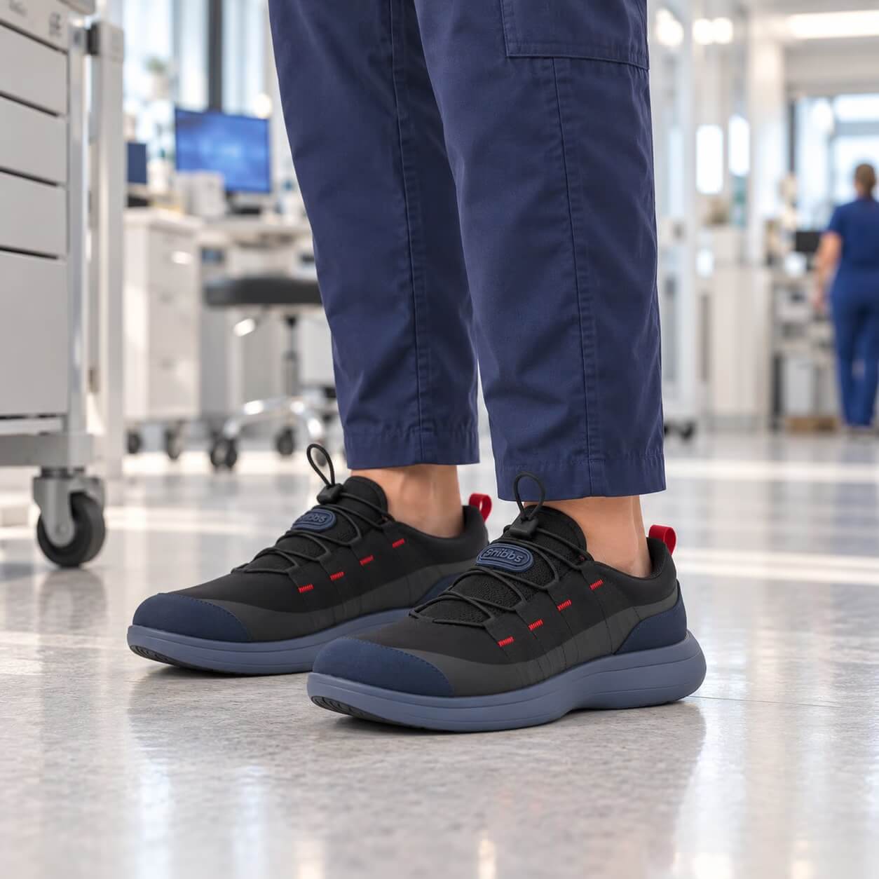 A close-up of a person wearing blue scrubs and black and blue sneakers in a hospital hallway.