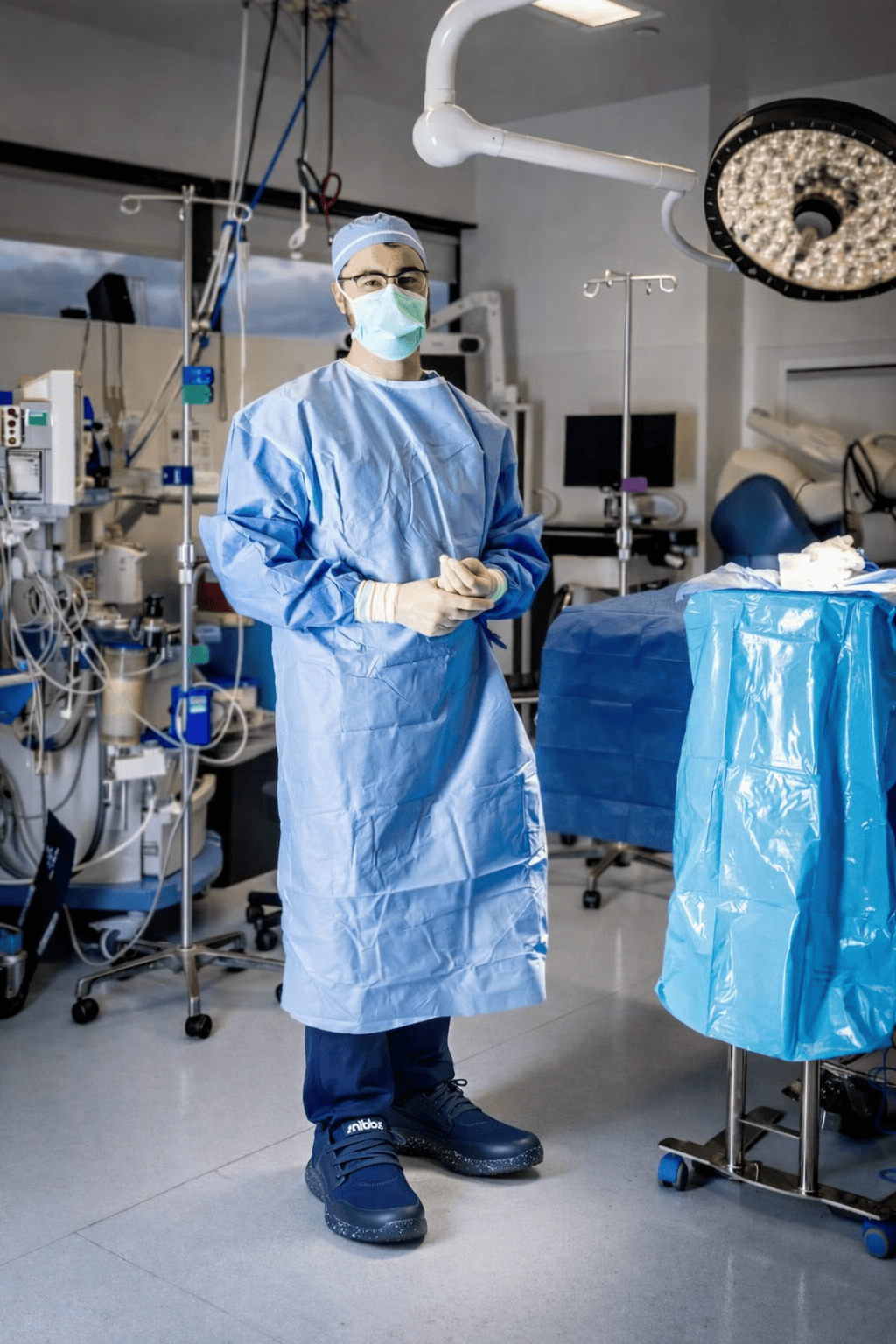 A surgeon in full surgical attire, including a mask and glasses, stands in an operating room.