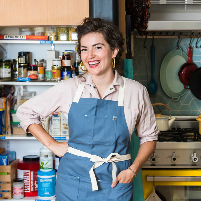 Person in a kitchen wearing a blue apron, smiling with shelves of ingredients behind.