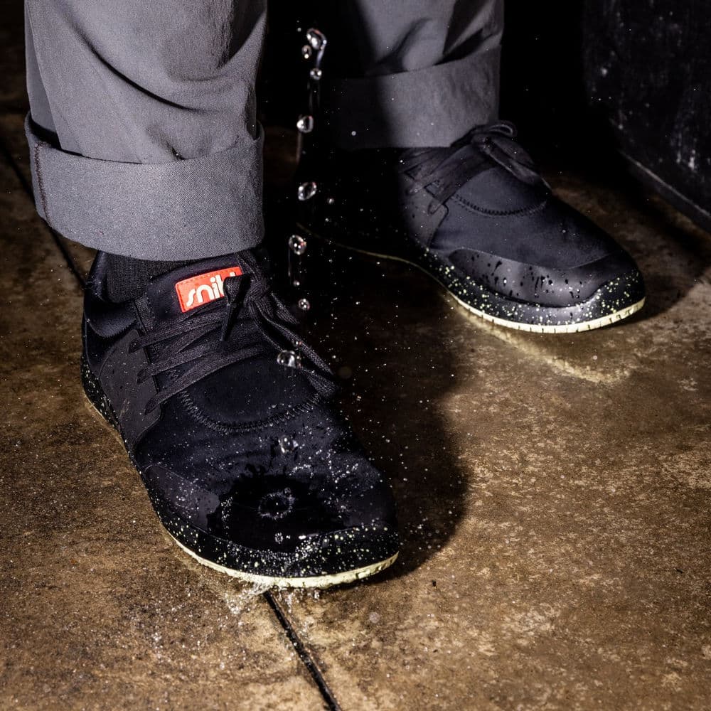 A close-up of water splashing onto a pair of black sneakers on a wet surface.