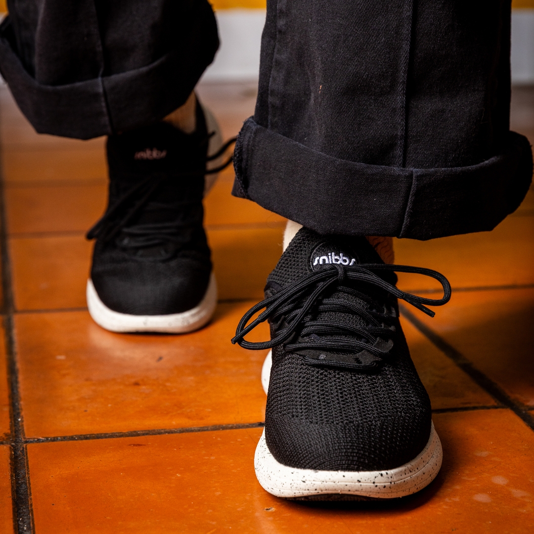 Person wearing black sneakers with white soles on a tiled floor.