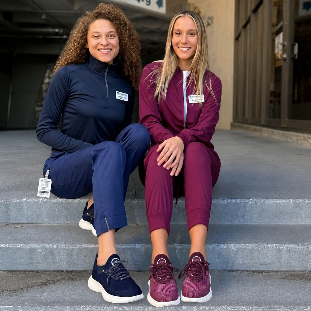 Two women in scrubs and athletic shoes sit smiling on concrete steps.
