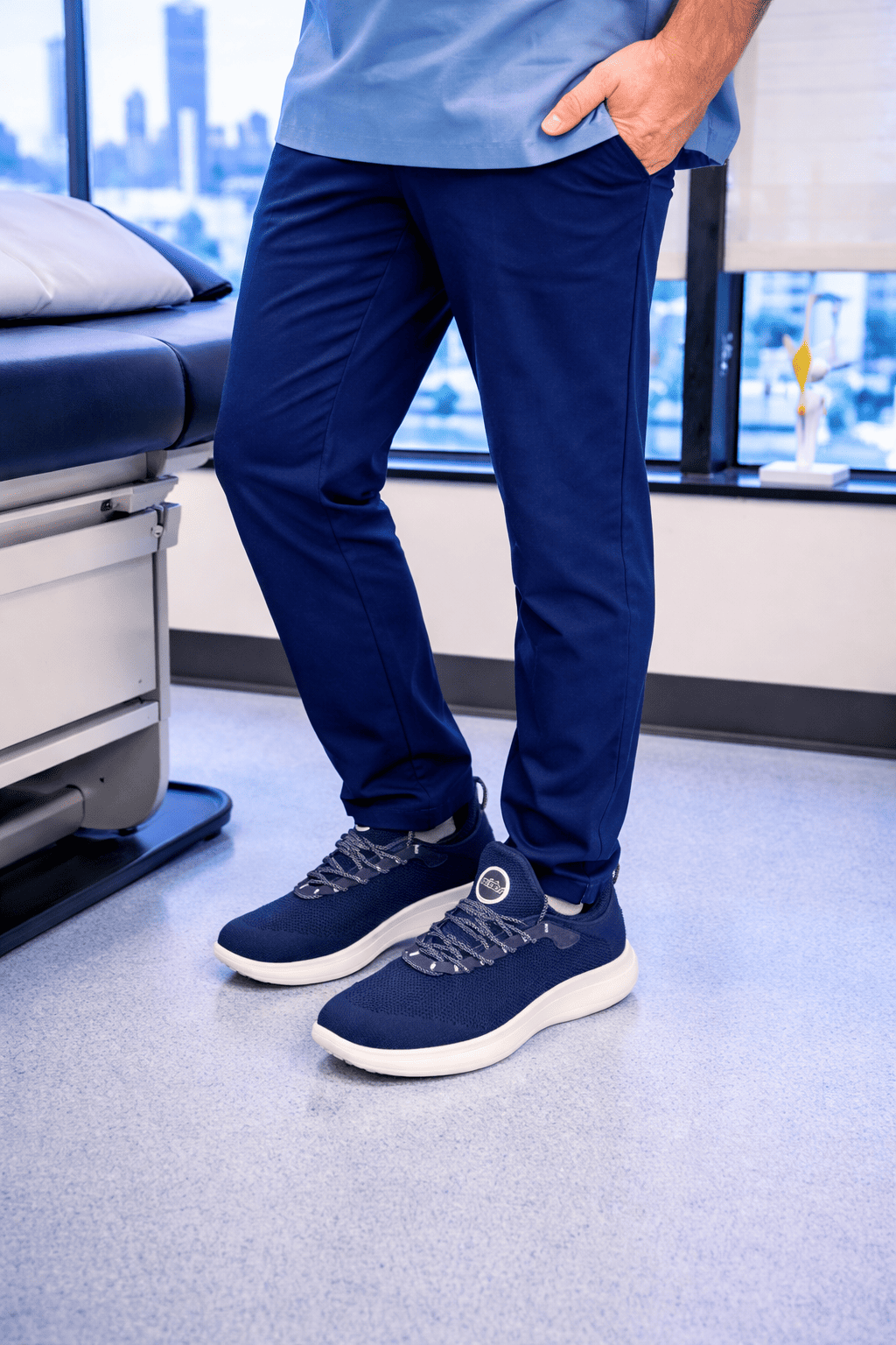 A person wearing blue scrubs and navy sneakers stands in a medical exam room.