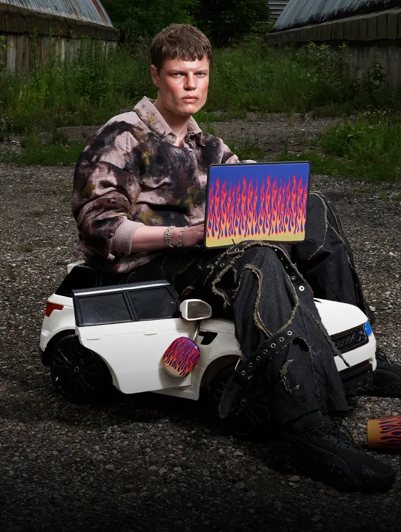A person sits on a white toy car while holding a laptop decorated with a flame pattern.