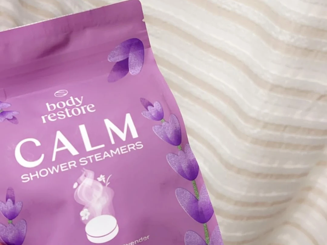 A close-up of a purple pouch of Body Restore Calm Shower Steamers resting on striped bedding.