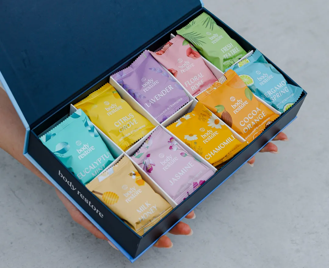 Hands holding an open blue gift box filled with an assortment of colorful Body Restore packets.