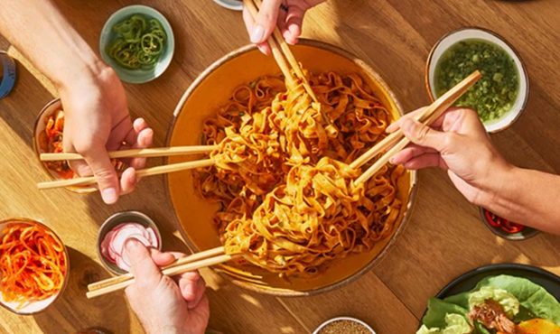 Four hands using chopsticks to eat noodles from a large bowl.