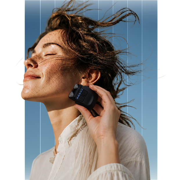 A woman with eyes closed holds a small electronic device to her jawline, her hair blowing in the wind.