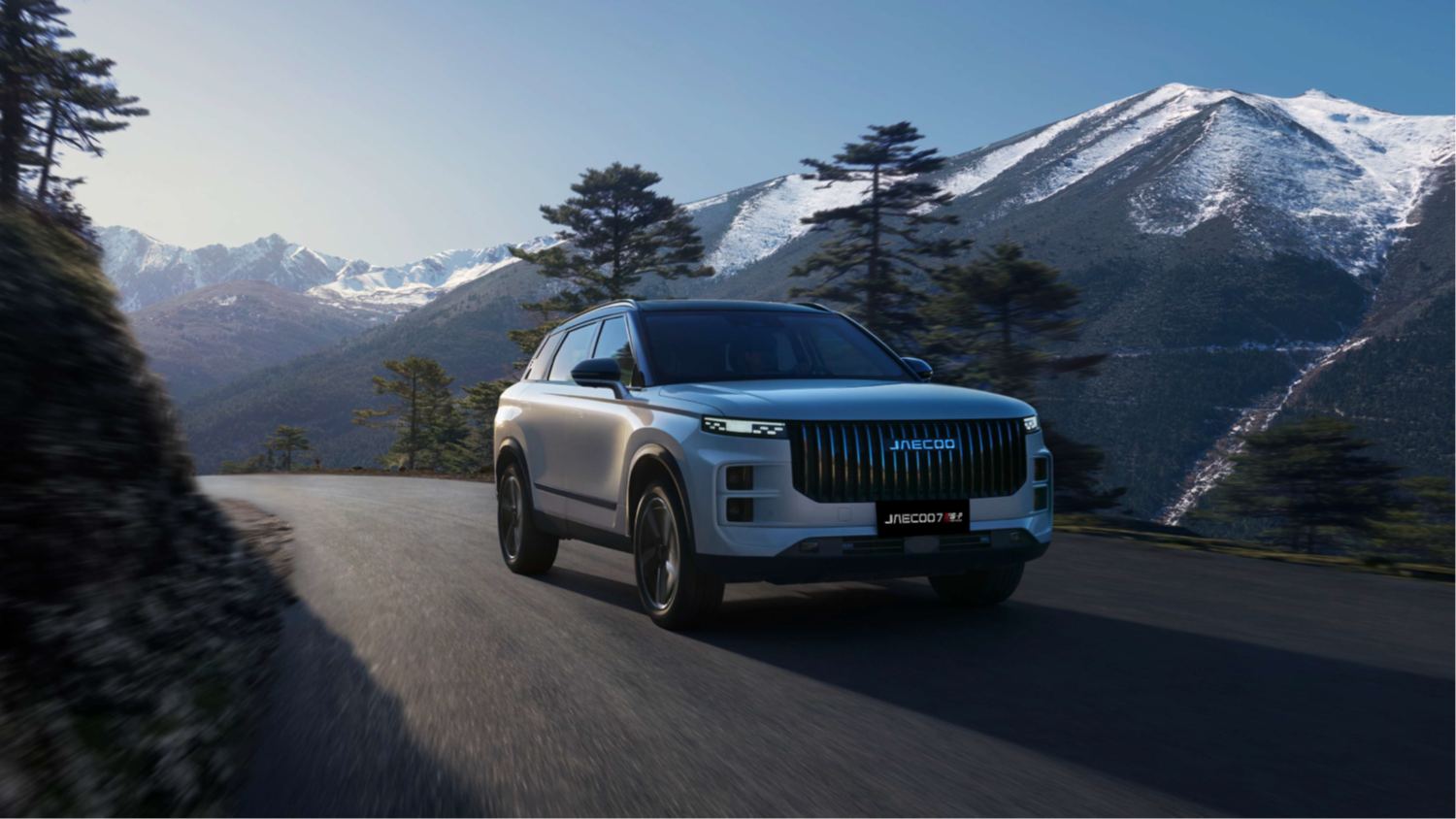 A silver Jaecoo SUV driving on a winding road with snow-capped mountains in the background.