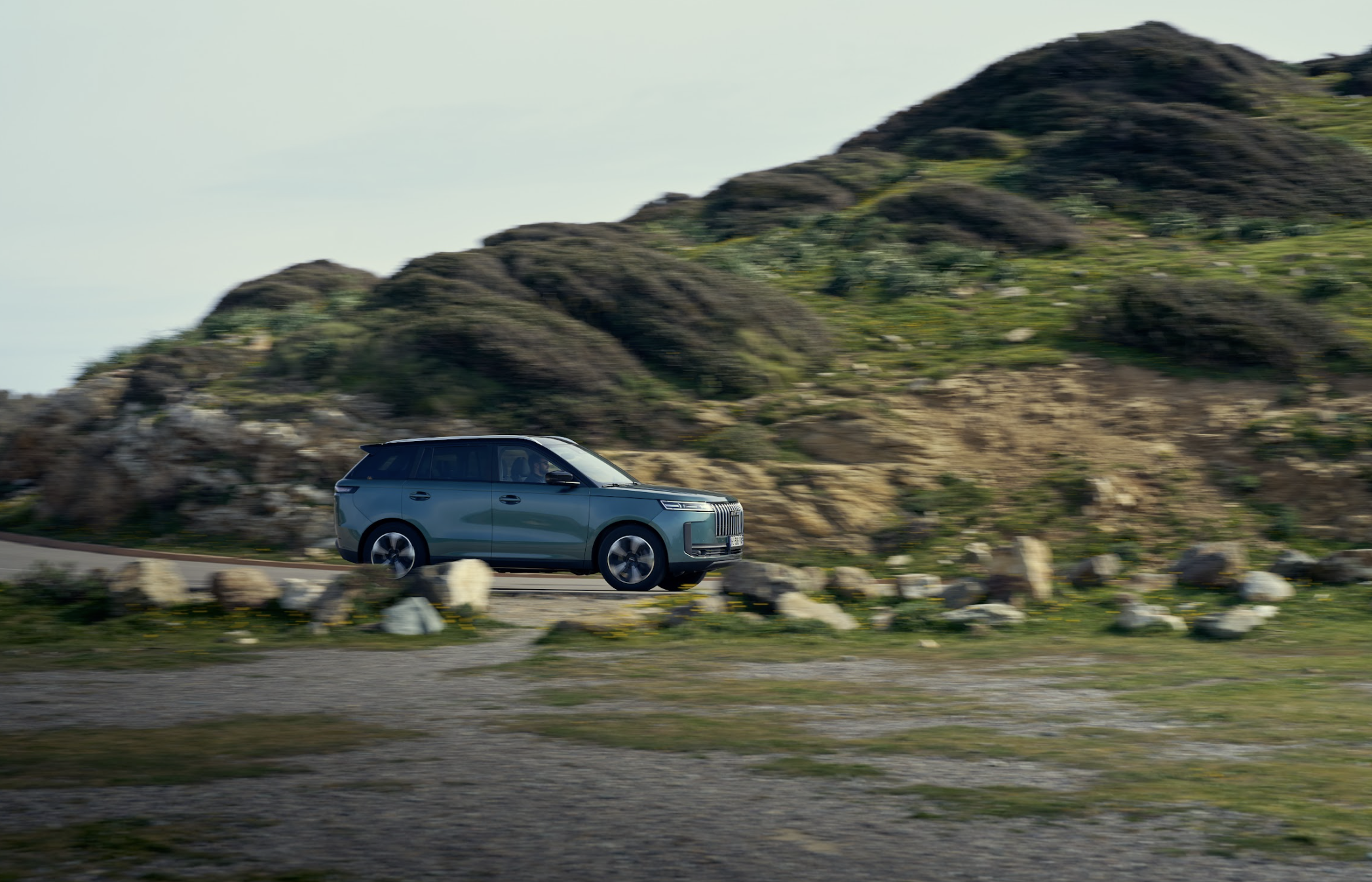 A blue-green SUV driving on a winding road with a motion-blurred background of a rocky, green hill.