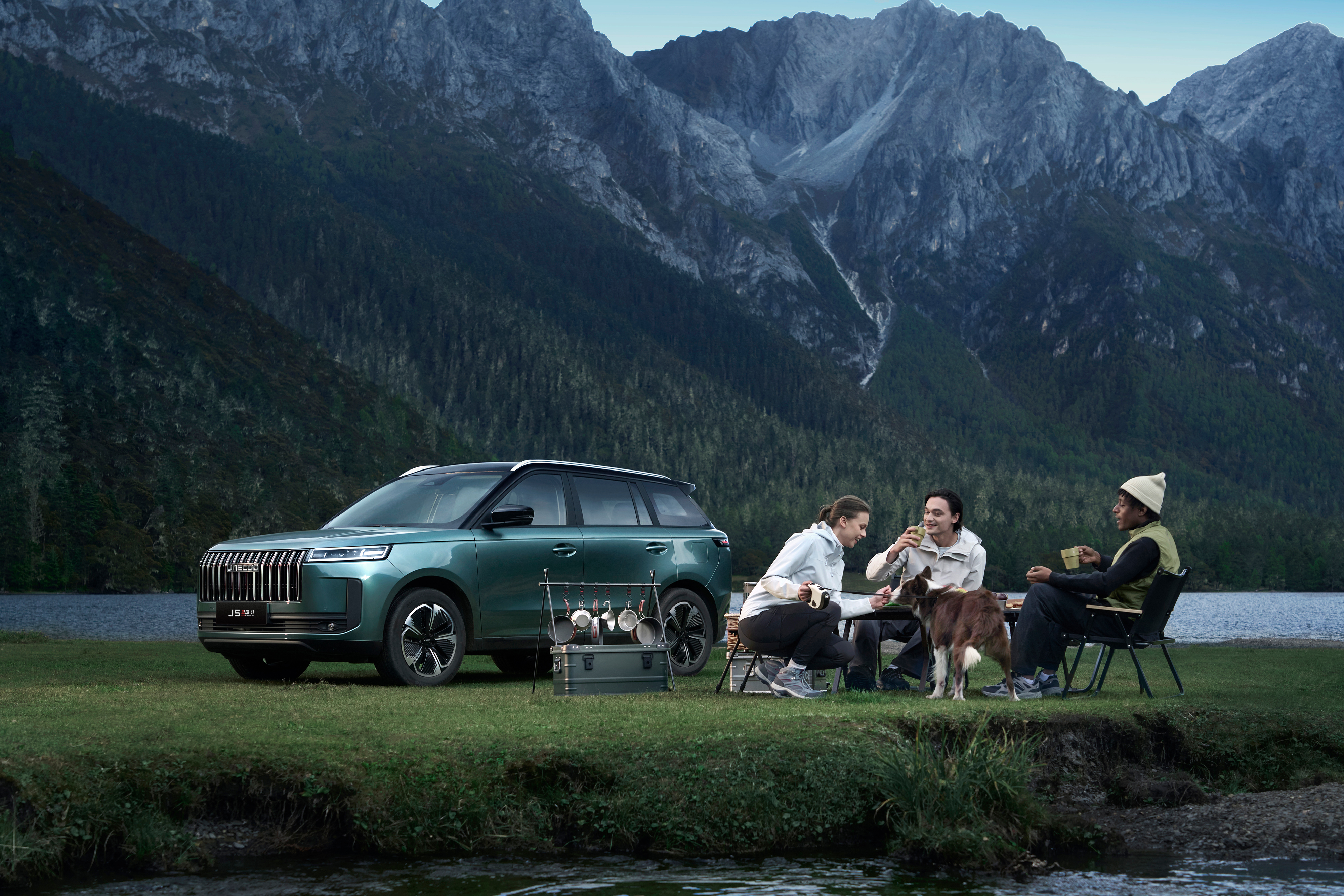 People sitting by an SUV near a lake with mountains in the background.
