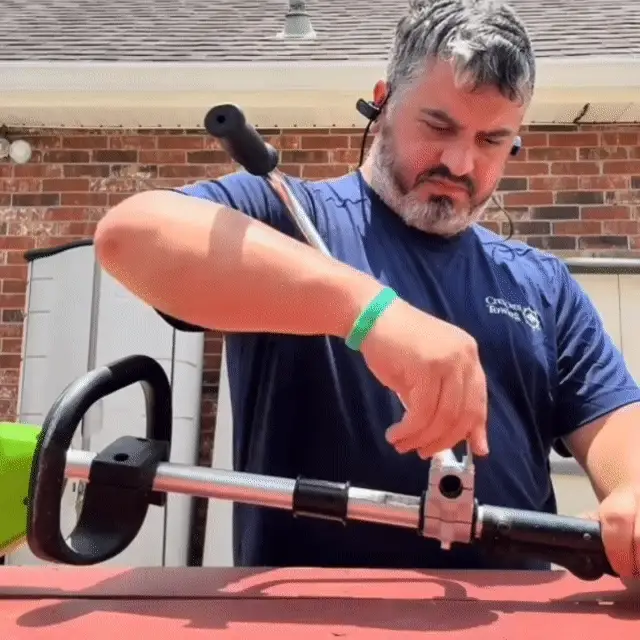 A man in a blue t-shirt and earbuds adjusts the clamp on a string trimmer outdoors.