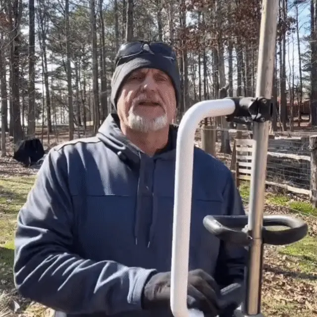 An older man in a beanie and jacket holds a piece of equipment while standing outdoors in a wooded area.