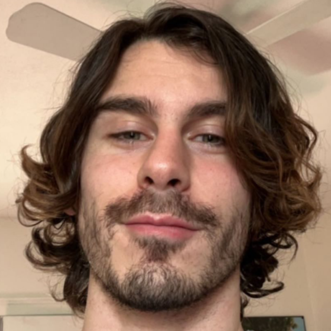 Person with wavy hair and facial hair, smiling slightly indoors.