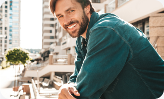 A smiling person in denim leaning on a railing outdoors in a city setting.